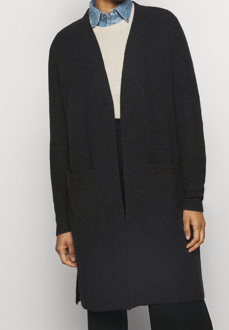 Dlouhý černý kardigan vyrobený z texturované pleteniny, s otevřeným předním dílem, dvěma předními kapsami a žebrovanými manžetami. Nosič je oblečený přes světlý svetr.