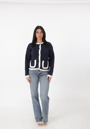 Cardigan lavorato a maglia navy con bordi bianchi, dotato di due grandi tasche frontali e scollatura rotonda, abbinato a jeans dritti blu chiaro.