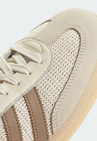 Gros plan sur une paire de sneakers crème et beige montrant des panneaux en tissu tissé, une cap toe en suède, une zone d'œillets en cuir, et une semelle en caoutchouc translucide texturée.