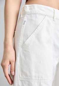 Pantalones de algodón blancos con bolsillos frontales en ángulo, una etiqueta de marca visible y detalles de costura. La tela parece suave con un acabado ligeramente texturizado.