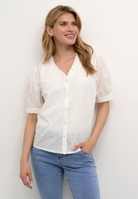 Camicia bianca con bottoni, maniche a sbuffo corte, motivo ad occhiello e scollatura ondulata. Indossata con jeans azzurri chiari.