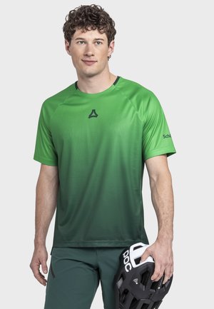 Junger Mann mit lockigem Haar, der ein grünes athletisches Shirt mit Farbverlauf und dunkle Shorts trägt, und einen schwarz-weißen Fahrradhelm in seiner rechten Hand hält.