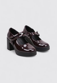 Par de zapatos Mary Jane de charol burdeos oscuro con doble correa, punta redonda y tacones bloque gruesos sobre un fondo gris claro.