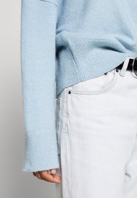 Jasnoniebieski sweter z dzianiny z szerokimi mankietami, w połączeniu z białymi dżinsami i czarnym paskiem, z widocznymi detalami metalowymi.