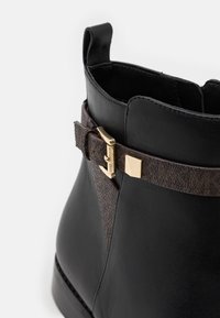 Bottine en cuir noir avec une bande marron texturée ornée de ferrures dorées et d'un détail logo. Le modèle comprend une languette pour un enfilage facile.