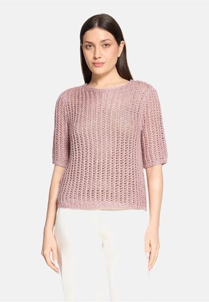 Frau mit langen dunklen Haaren, die einen hellrosa, locker gestrickten Kurzarmpullover und eine weiße Hose trägt, nach vorne blickend mit neutralem Gesichtsausdruck.