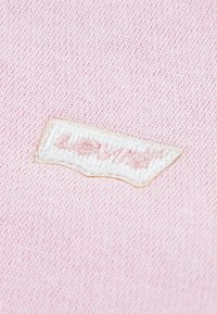 Rosa Baumwollstoff mit einem strukturierten weißen Etikett, auf dem "Levi's" in rosa Faden gestickt ist. Einfaches, rechteckiges Design mit weicher Textur.