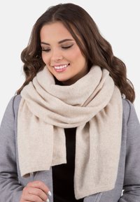 Écharpe en laine beige, texture douce, enroulée lâchement autour du cou, avec un design épuré et simple ; une partie d'un manteau gris visible en arrière-plan.