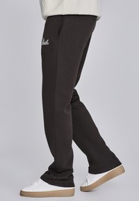 Donkergrijze joggingsbroek met zijzakken, elastische tailleband en een witte geborduurde logo. Gepaarde met witte sneakers met beige zolen.