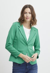 Zelený blazer vyrobený z měkké tkaniny, s klopovým límcem, zapínáním na jeden knoflík a dvěma předními kapsami. Nosený přes bílý top.