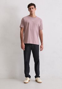 Młody mężczyzna stoi przy białej ścianie, ubrany w fioletową koszulę z krótkim rękawem, ciemne spodnie i białe trampki z gumowymi podeszwami.