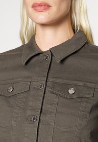 Grön denimskjorta med krage, som har två bröstfickor och metallknappar. Tyget har ett slätt, strukturerat utseende.