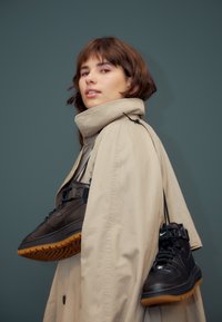 Schwarze High-Top-Sneaker mit Gummisohlen, aufgehängt an schwarzen Riemen. Das Modell trägt einen beigen Trenchcoat und zeigt ein modernes, minimalistisches Design.