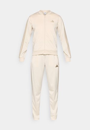 Kremowy dres Adidas z pełnym zamkiem błyskawicznym, bocznymi kieszeniami, ściągaczami u mankietów oraz pasujące spodnie z logo na klatce piersiowej i udzie.