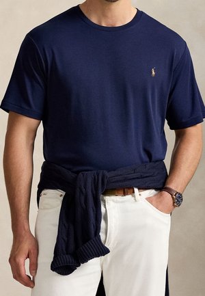 Mann trägt dunkelblaues T-Shirt mit kleinem Logo, weiße Hose, braunen Gürtel, dunkelblauen Pullover um die Taille gebunden und Armbanduhr am linken Handgelenk.