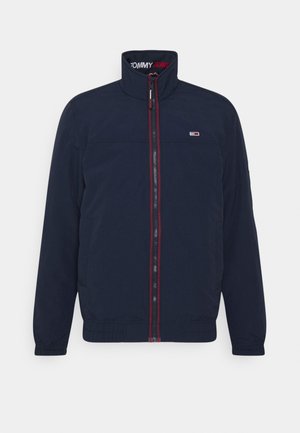 Marineblaue Jacke zum Reißverschluss mit hohem Kragen, rotem Reißverschlussbesatz, elastischen Bündchen und kleinem Tommy Jeans-Logo auf der Brust und im Krageninnenbereich.