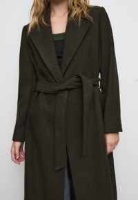 Cappotto lungo di un verde scuro con cintura in vita, ampi rever e maniche lunghe; realizzato in un tessuto morbido e strutturato; apertura frontale con taglio dritto.