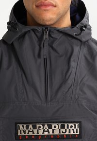 Grå pulloverjakke med hætte, der har en lynlås foran og et markant sort og sølv logo på brystet. Glat, mat tekstur.