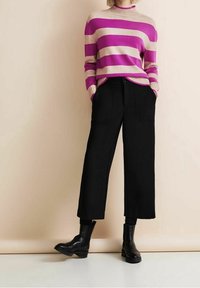 Person som bär en randig tröja i rosa och beige, svarta korta vida byxor och svarta ankelstövlar, står med händerna i fickorna.