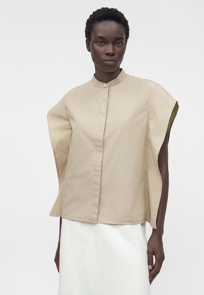 Άτομο με σκούρο δέρμα φοράει μπεζ, φαρδιά αμάνικη μπλούζα με ψηλό γιακά και λευκή φούστα, στέκεται μπροστά σε ένα απλό γκρι φόντο.