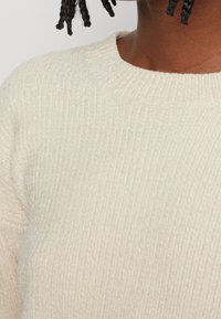 Person mit dunklem, geflochtenem Haar, der einen cremefarbenen, gerippten, strukturierten Strickpullover mit rundem Ausschnitt vor einem neutralen Hintergrund trägt.