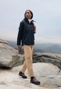 Black waterproof jacket with a hood, beige cargo pants, and black hiking shoes with pink laces, on rocky terrain under a gray sky.
