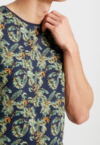 Man in een donkerblauw shirt met een groen tropisch blad en een oranje bloemenpatroon, hand omhoog bij de nek tegen een witte achtergrond.