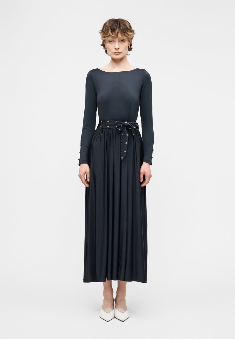 Γυναίκα όρθια που φοράει μαύρο midi φόρεμα με μακριά μανίκια και ζώνη στη μέση, καθώς και λευκά παπούτσια με μυτερή μύτη, σε λευκό φόντο.