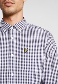 Camicia a quadri vichy in cotone navy e bianco, con una piccola tasca e un logo giallo ricamato sul lato sinistro del petto. Collo button-down.