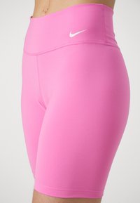 Shorts de sport roses en matériau extensible, avec une taille haute et un petit logo blanc sur le côté. Texture lisse, coupe ajustée.