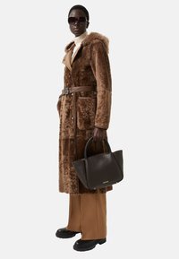 Hnědý ovčí kabát s kapucí, předními knoflíky a hnědým koženým opaskem. Hnědá kabelka s dvojitými uchy; model má na sobě tmavé sluneční brýle a kalhoty.