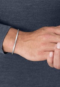 Brazalete de metal en una muñeca; forma redonda y elegante con acabado mate en color plata; llevado sobre una manga de punto gris. Textura suave, diseño minimalista.