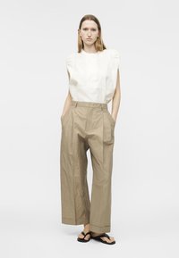 Γυναίκα που φοράει λευκό αμάνικο τοπ, χαλαρά μπεζ παντελόνια με φαρδύ πόδι και μαύρα σανδάλια, στέκεται με τα χέρια στις τσέπες σε λευκό φόντο.