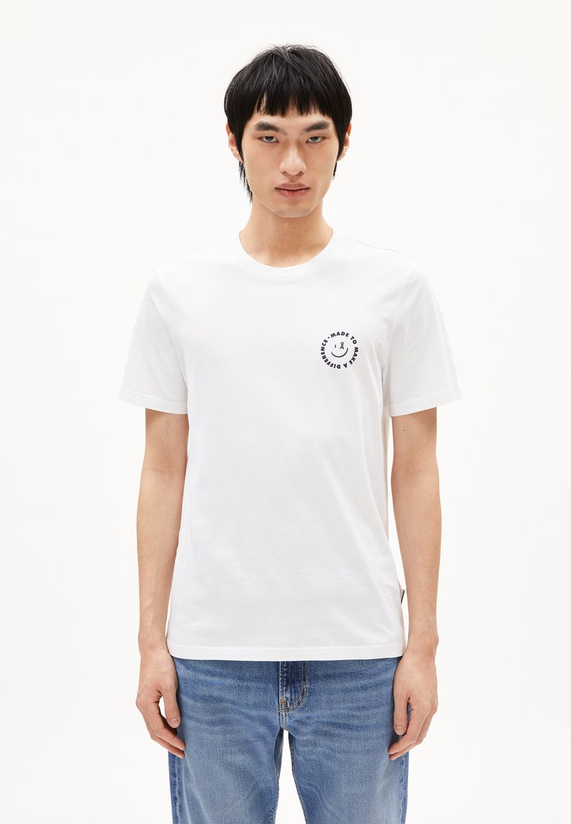 Weißes Baumwoll-T-Shirt mit Rundhalsausschnitt, kurzen Ärmeln und einem schwarzen kreisförmigen Logo auf der linken Brust. Getragen mit blauen Jeans.