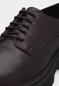 Σκούρο καφέ οξφορντ δερμάτινο παπούτσι με μαύρα στρογγυλά κορδόνια και παχιά μαύρη καουτσούκ σόλα, εμφανιζόμενο σε κοντινή λήψη σε λευκό φόντο.