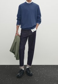 Pull en maille marine, chemise blanche à col, jean foncé, chaussettes grises retroussées, chaussures noires et un pull côtelé vert posé sur un bras.