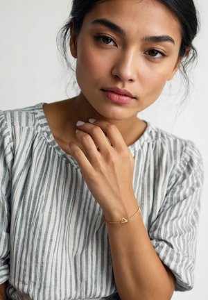 Gyldent armbånd med en trekantet charm på en delikat kæde, båret på en hånd med neutralt neglelak. Model iført en grå stribet skjorte.