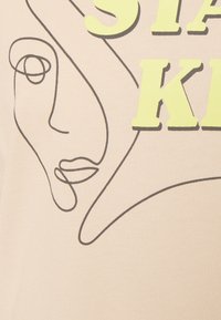 Camiseta de tela beige con un dibujo de línea negra de un rostro y un texto en amarillo negrita que dice "STAY KID". Textura suave con un diseño minimalista.