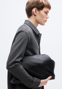 Μαύρη δερμάτινη τσάντα που κρατιέται κάτω από το χέρι, με απαλή υφή, μινιμαλιστικό σχέδιο. Γκρι παλτό με υφή και γιακά, πλαϊνή όψη ενός ατόμου.