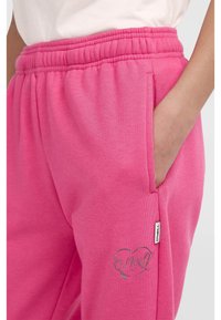 Roza trenerka z elastičnim pasom in stranskimi žepi. Ima majhen srebrn logotip na nogi in mehko bombažno teksturo.