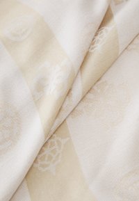 Tekstil dalam warna krim dan beige terang dengan pola halus, termasuk kerang dan bentuk geometri, menampilkan tekstur tenun yang lembut.