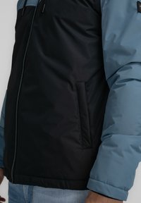 Chaqueta acolchada con una sección superior azul y una sección inferior negra, cierre frontal con cremallera, bolsillo lateral y puños acanalados para mayor calidez.