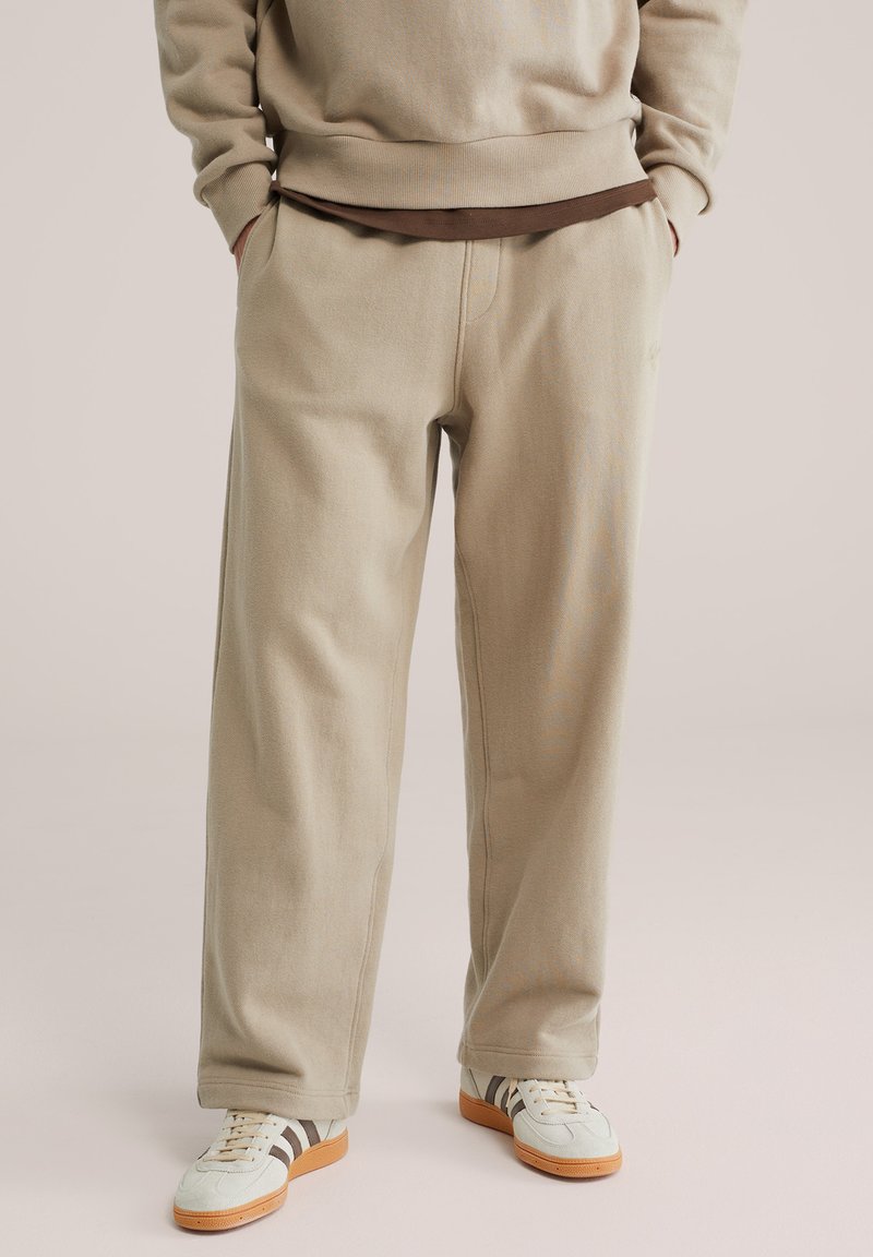 Osoba nosząca beżowe spodnie o szerokich nogawkach, beżową bluzę nałożoną na brązową koszulę oraz białe trampki z brązowymi paskami i gumowymi podeszwami