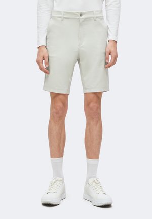 Personne portant un short gris clair arrivant au genou, une chemise blanche à manches longues, des chaussettes blanches et des baskets blanches, debout devant un fond uni.