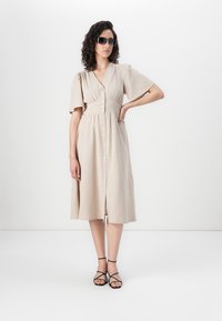 Abito midi beige con un davanti a bottoni, maniche corte a palloncino e vita aderente. Il modello indossa occhiali da sole neri e sandali con cinturini.