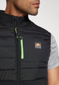 Czarna puchowa kamizelka z zamkiem błyskawicznym z przodu, zielonym akcentem na suwaku i logo ellesse. Posiada wysoki kołnierz i pikowaną fakturę.
