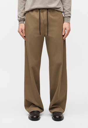 Personne portant un pantalon ample à jambes larges marron avec un cordon noir, associé à des chaussures marron foncé et un pull beige clair.