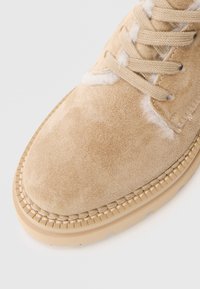 Chaussure en suède beige avec une finition texturée, dotée de lacets, d'accents en fourrure et d'une semelle en caoutchouc léger avec des coutures visibles.