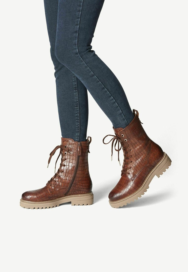 Bottines marron à lacets avec fermeture éclair latérale et motif texturé portées avec un jean skinny foncé sur fond blanc.