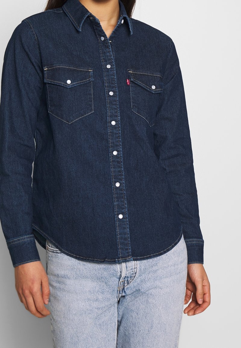 Personne portant une chemise en denim bleu foncé avec des boutons-pression blancs et deux poches poitrine boutonnées, associée à un jean bleu clair.